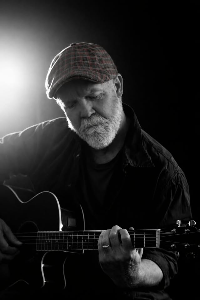 Older man with a plaid cap playing an acoustic guitar, eyes closed, in a moody black-and-white portrait.