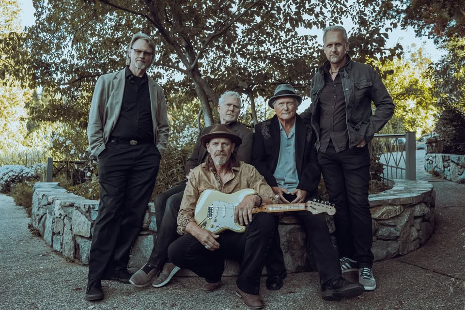 Five-man rock band posing outdoors on a stone wall, with the guitarist seated in front holding a white electric guitar.
