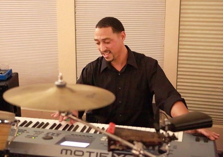 Man playing a MED keyboard and drum set, smiling while using both hands at the keys and cymbal nearby, indoor studio.