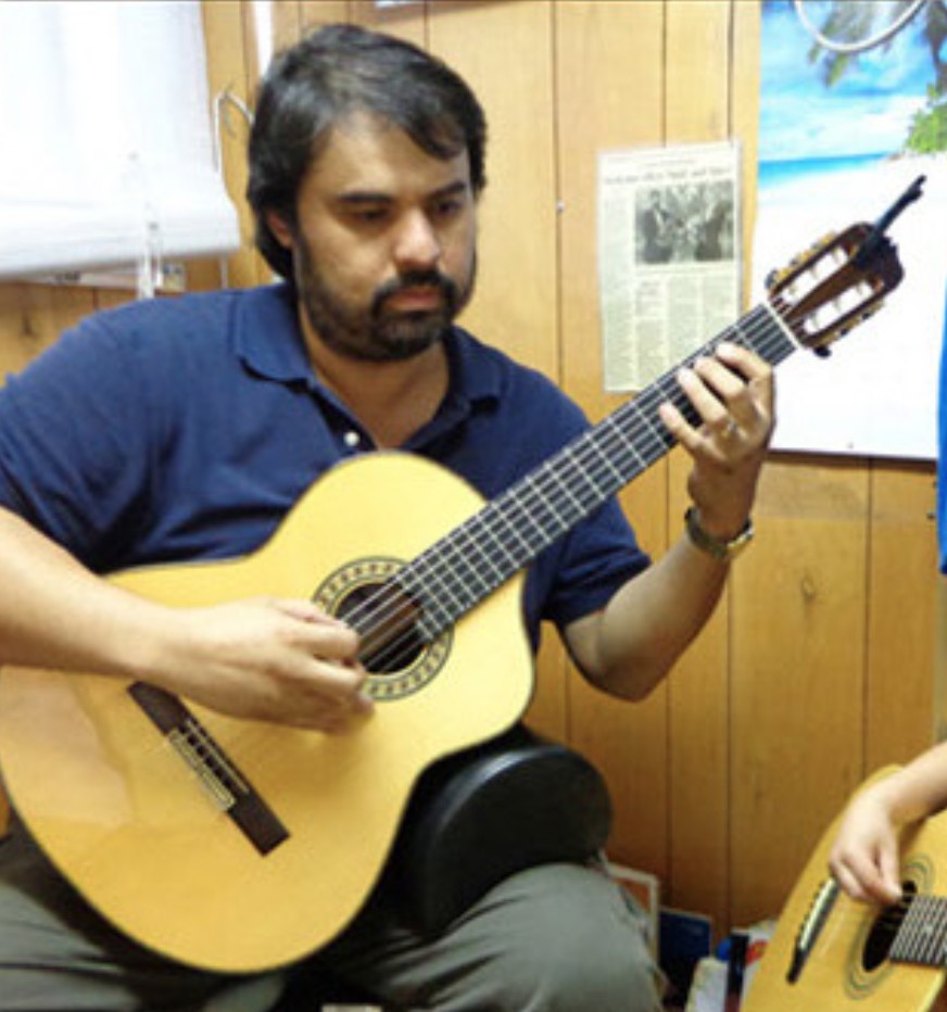 Man plays a classical guitar indoors, focusing on the strings, with another guitar visible nearby.