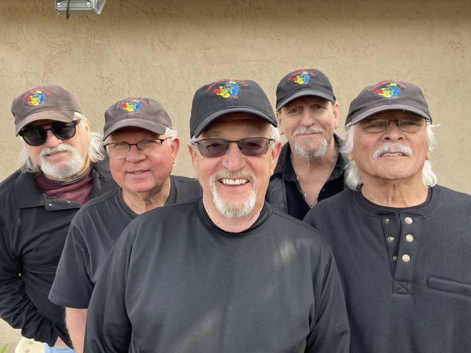 Five men in black shirts and matching caps with a colorful crest, posing and smiling against a beige wall.