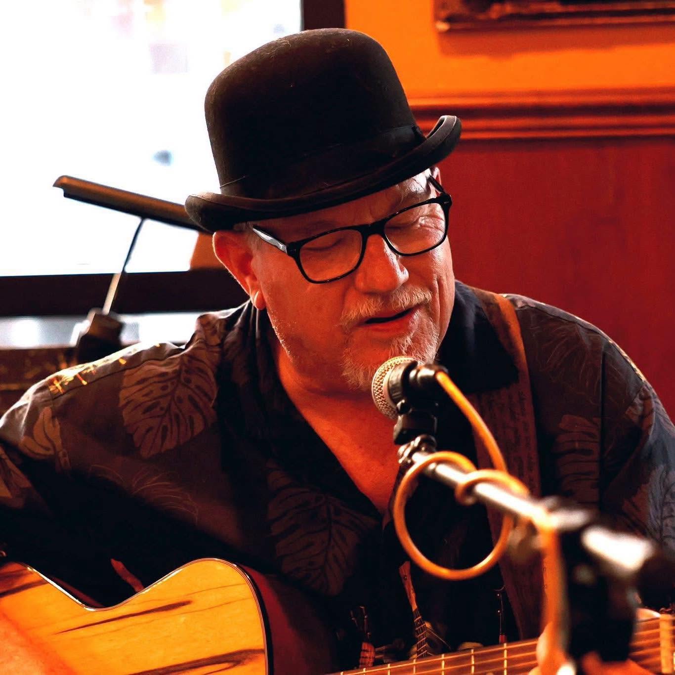 Male musician wearing glasses and a hat, singing into a microphone while playing an acoustic guitar at a live performance