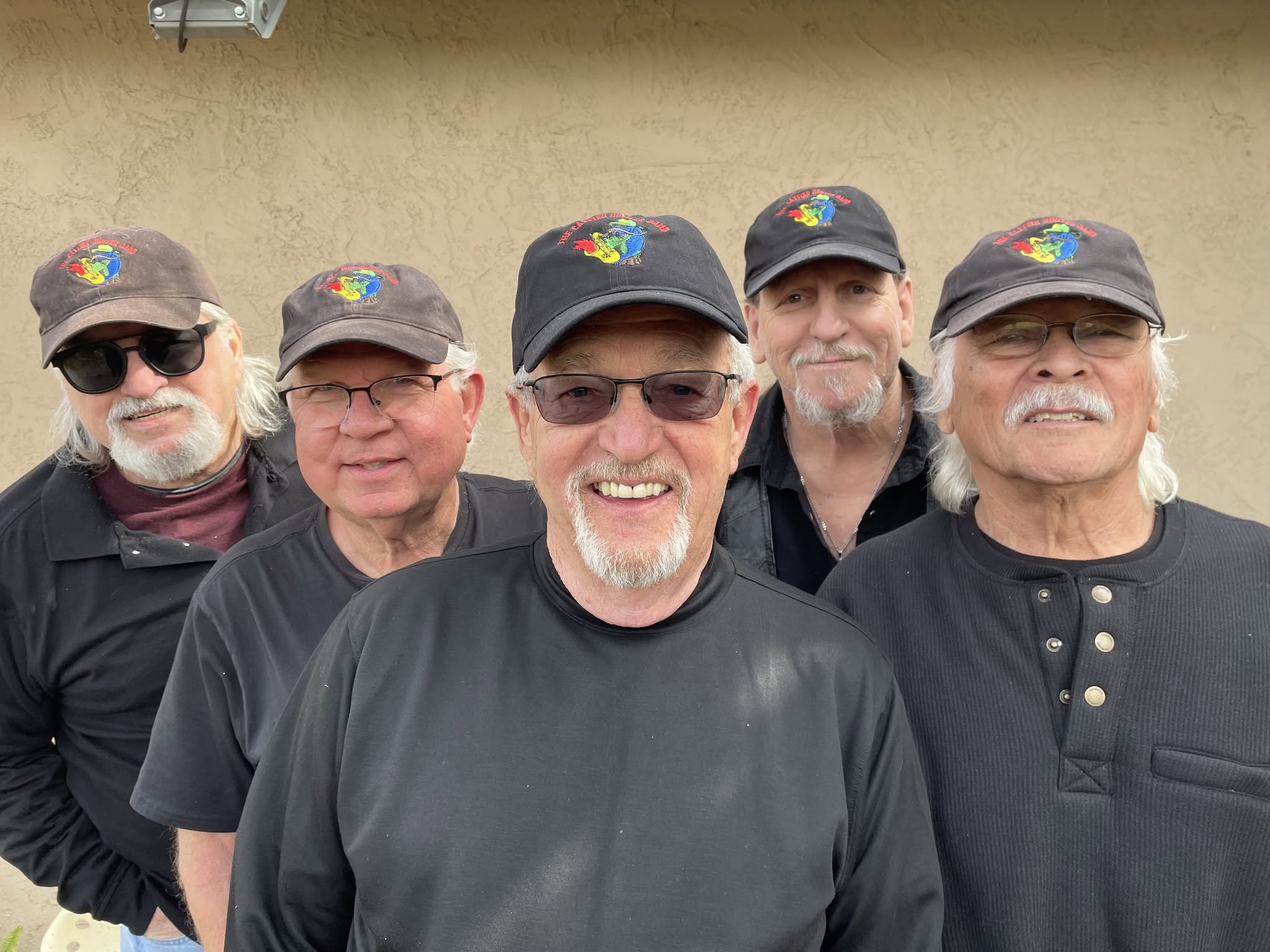 Group of five men wearing black shirts and matching hats with a colorful logo, posing for a selfie outdoors.