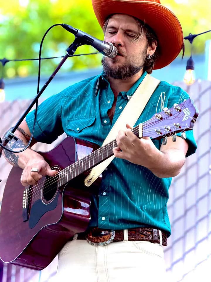Male musician wearing an orange cowboy hat and teal shirt plays a purple acoustic guitar into a microphone on an outdoor stage.
