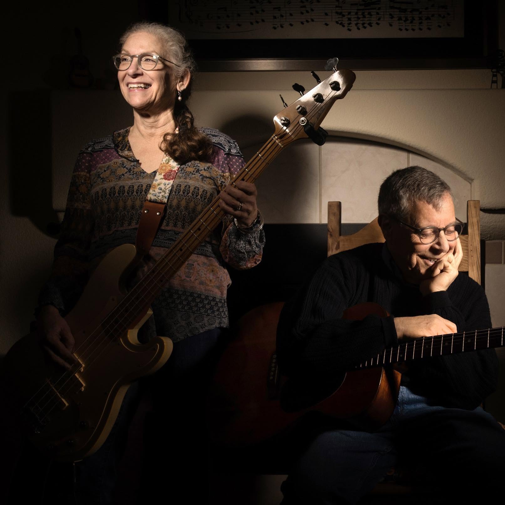 Smiling woman with glasses playing a bass guitar, seated beside a man who rests a hand on his guitar in a dimly lit room by a fireplace.