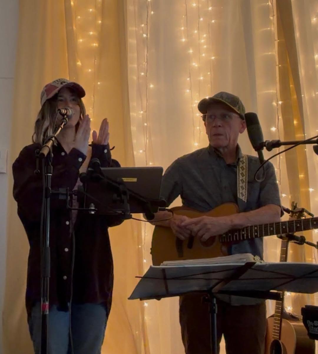 Two musicians performing on stage: a woman singing into a microphone and clapping, beside a man with an acoustic guitar under warm string lights.