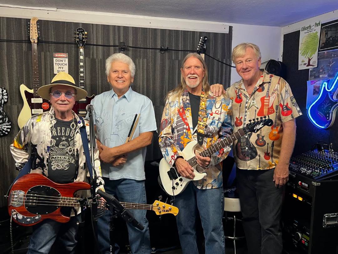 Four older men posing with guitars in a music studio, smiling for the camera, with amps and a neon guitar sign in the background.