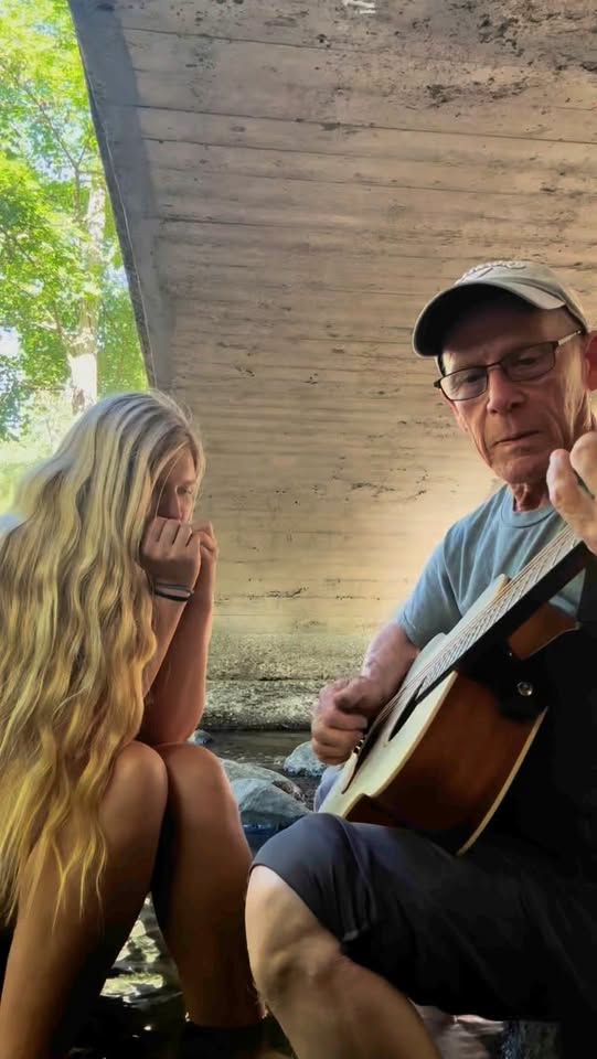 Older man in a cap plays an acoustic guitar under a concrete bridge while a young woman with long blonde hair watches nearby.