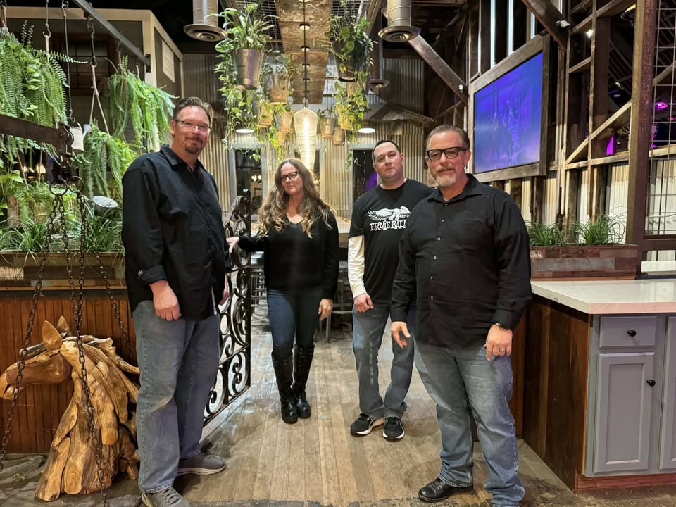 Four adults stand together in a plant-filled, rustic indoor space, posing for a group photo.